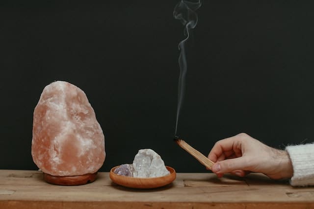 A hand burning a wooden stick next to a crystal and Himalayan salt lamp